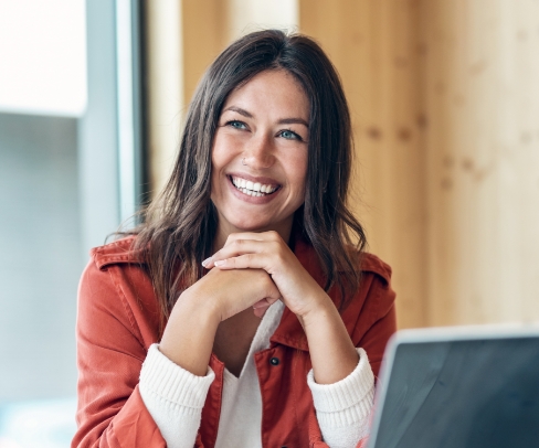Frau sitzt vor einem laptop uns stützt sich auf die arme, lächelt in die Kamera